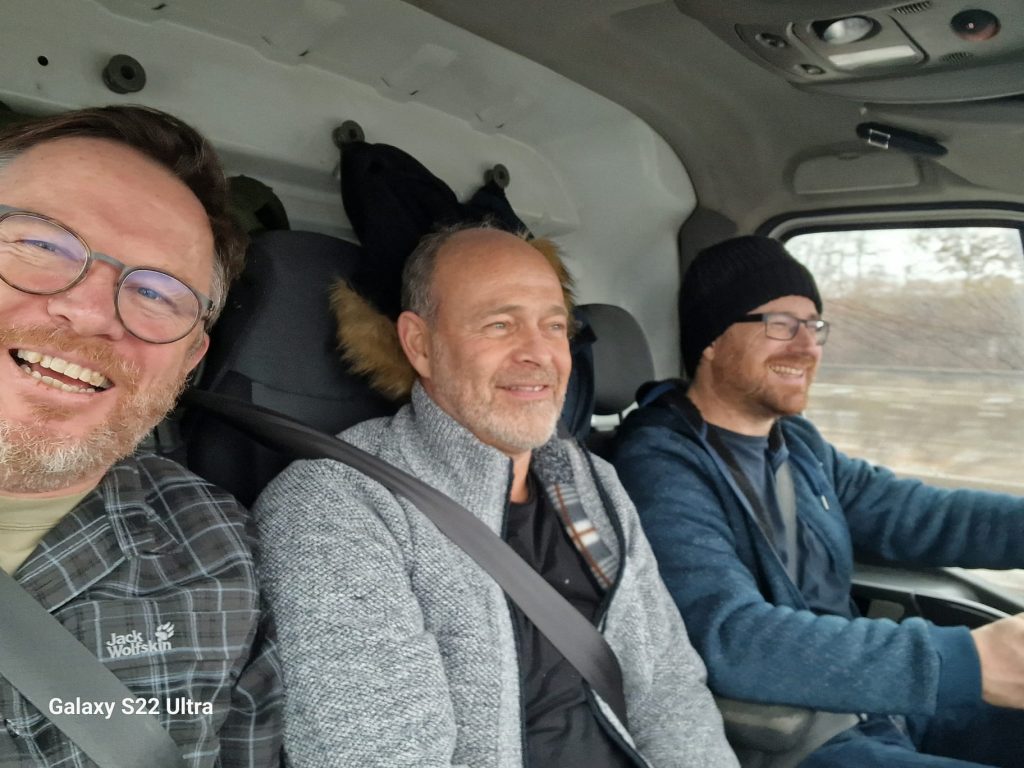 Zu sehen isind die drei Kollegen im Transporter, auf dem Weg in die Ukraine. Sie lachen in die Kamera.
